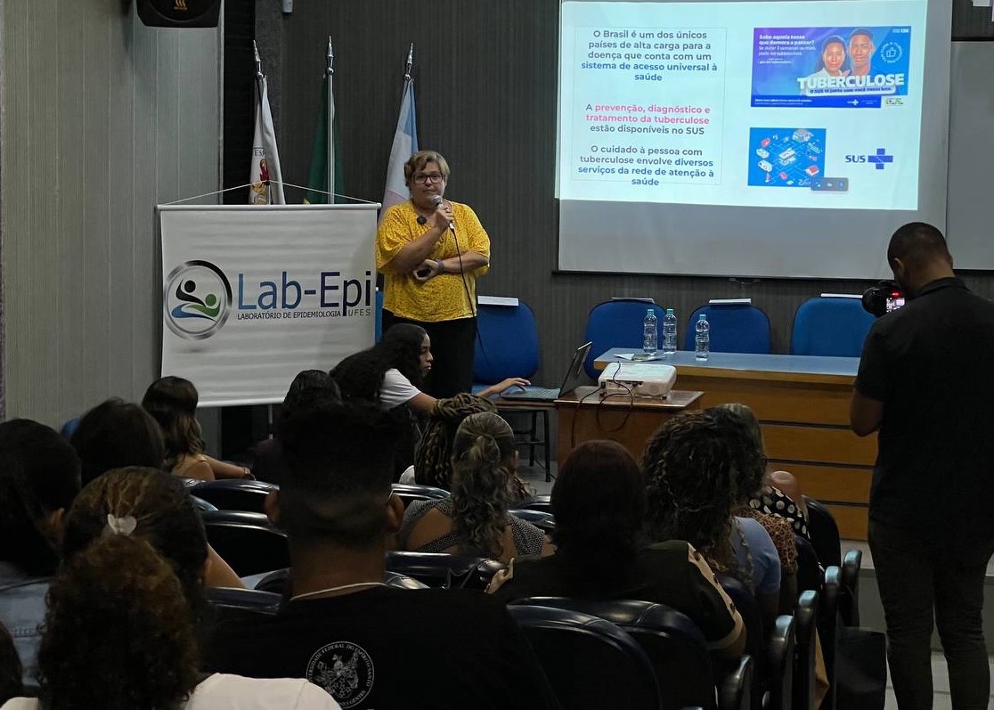 Foto do público do evento dentro do auditório, assistindo à fala de Fernanda Dockhorn, à frente, de camisa amarela.