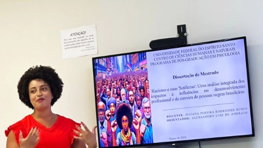 Foto da pesquisadora Juliana Nunes, em pé, apresentando sua pesquisa. Ela é uma mulher negra, de cabelos cursos e veste vestido vermelho.