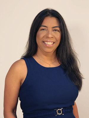 Fotografia de mulher de pele marron e cabelos longos pretos, sorrindo e usando blusa azul escura. fundo liso bege