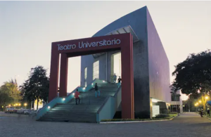 teatro da Ufes - prédio na cor cinza com a parede lateral esquerda arredondada e um portal grande vermelho na frente e tendo abaixo do portal uma escadaria