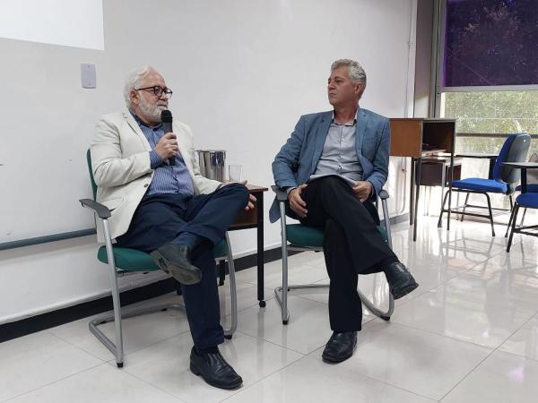 Reitor e vice-reitor da Ufes sentados em cadeiras, com o reitor falando ao microfone e o vice-reitor olhando para ele