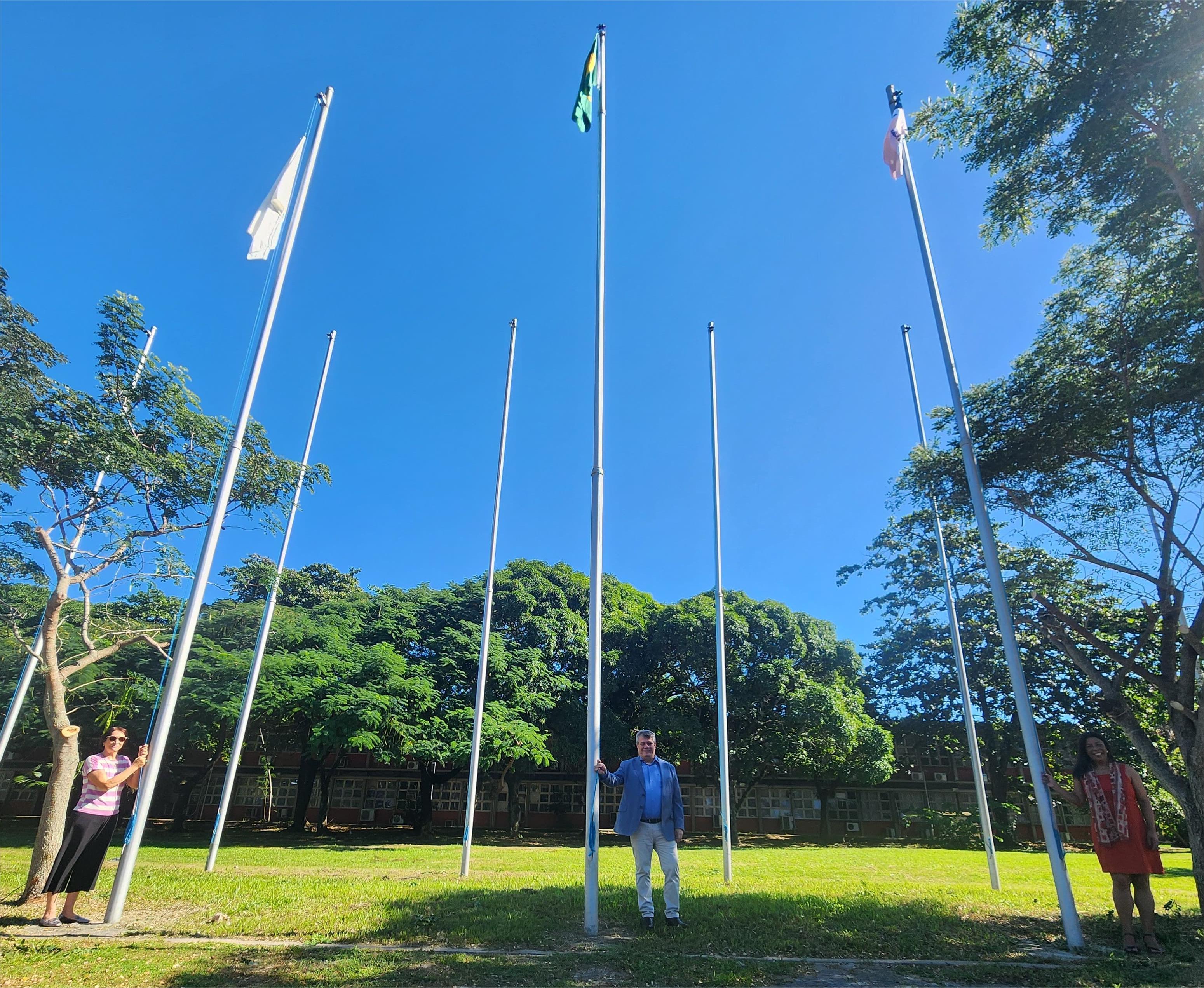 A servidora Santinha Girardi, o reitor Eustáquio de Castro e a vice-reitora Sonia Lopes posam em frente aos mastros das bandeiras. 
