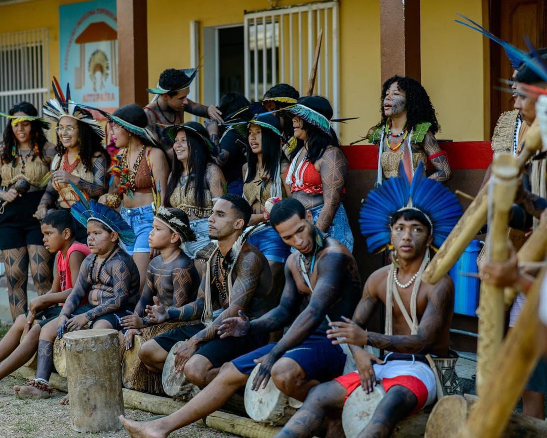 Grupo de dança Jovem Tupinikim, de Aracruz - ES. A foto mostra um grupo de jovens indígenas com os corpos pintados usando cocares e  acessórios indígenas. Alguns tocam pequenos tambores.