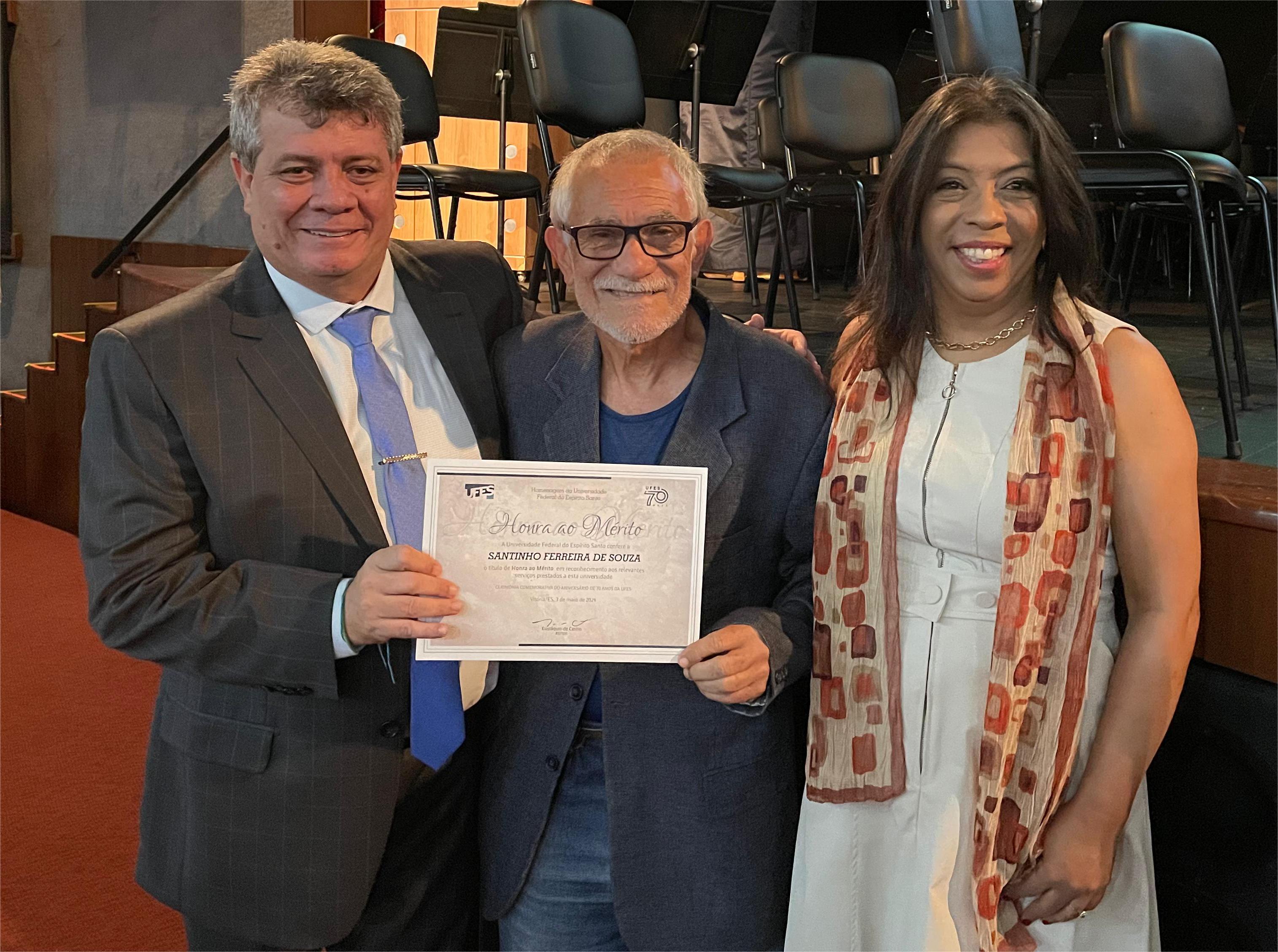 O reitor e a vice-reitora ladeiam o professor homenageado, Santinho de Souza, que segura o título de honra ao mérito