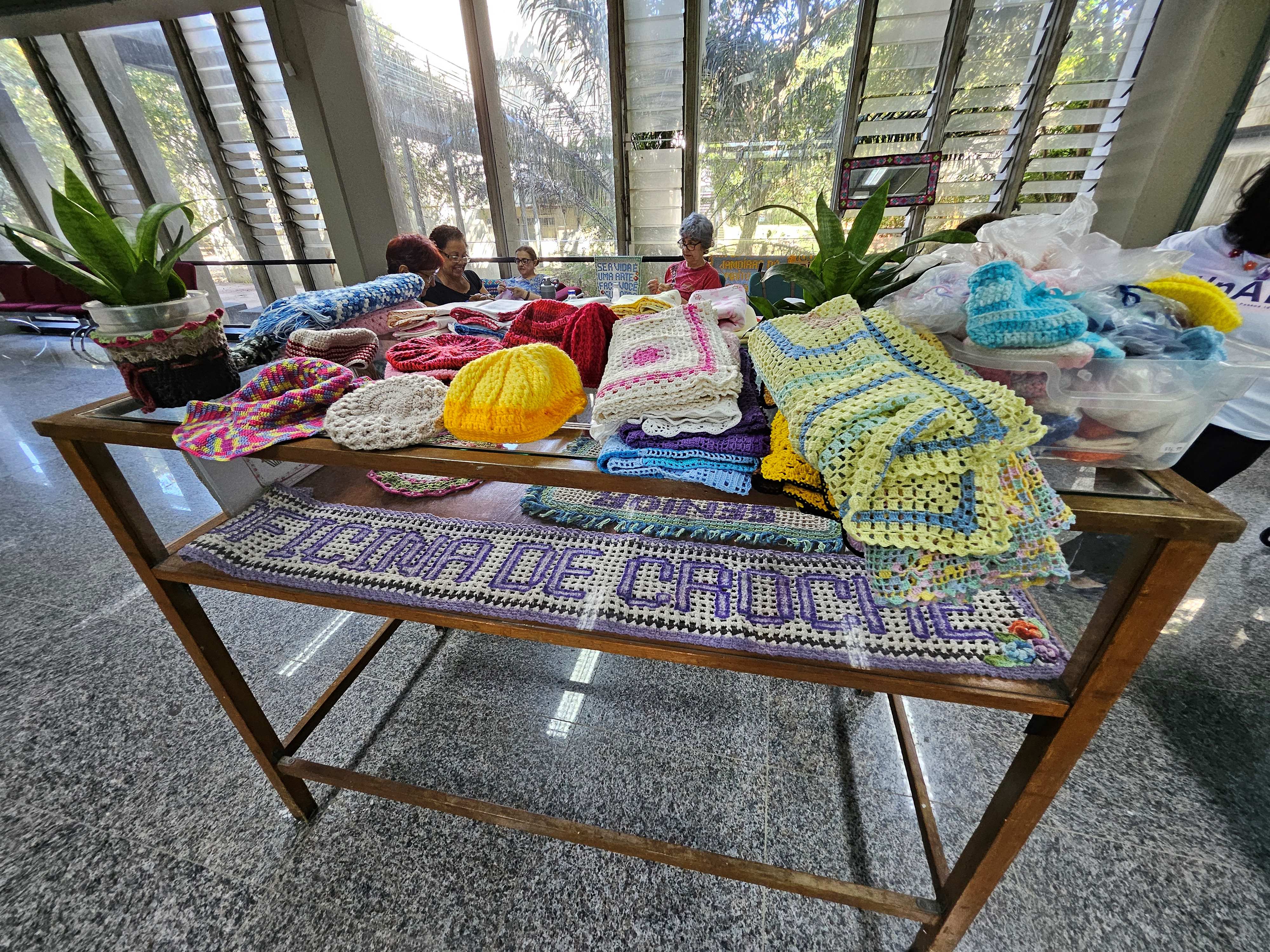 Mesa com as peças de crochê produzidas na oficina. Ao fundo, as participantes crochetando