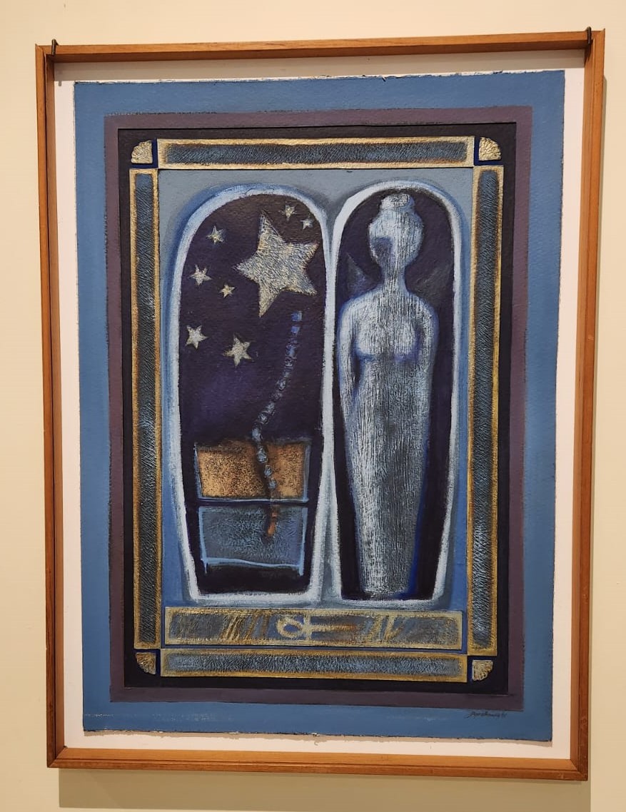 Foto de um dos quadros da exposição. A imagem mostra um retângulo azul dividido ao meio. De um lado, duas caixas sobrepostas de onde sai um cordão que leva a estrelas. Do outro, a escultura da silhueta de uma mulher.