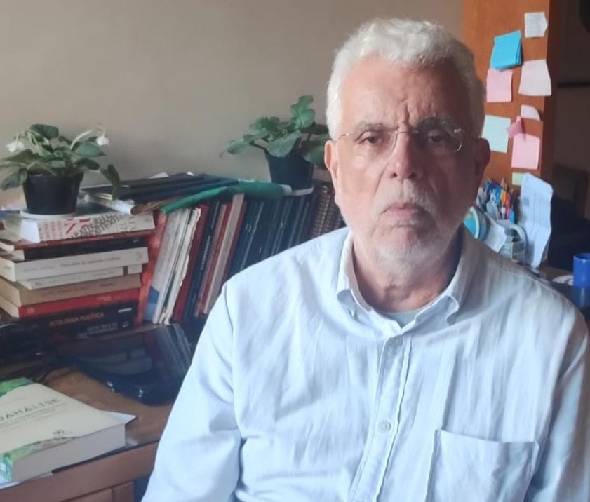 Foto do professor Roberto Simões sentado, vestindo uma camisa branca, ao lado de uma mesa com alguns livros