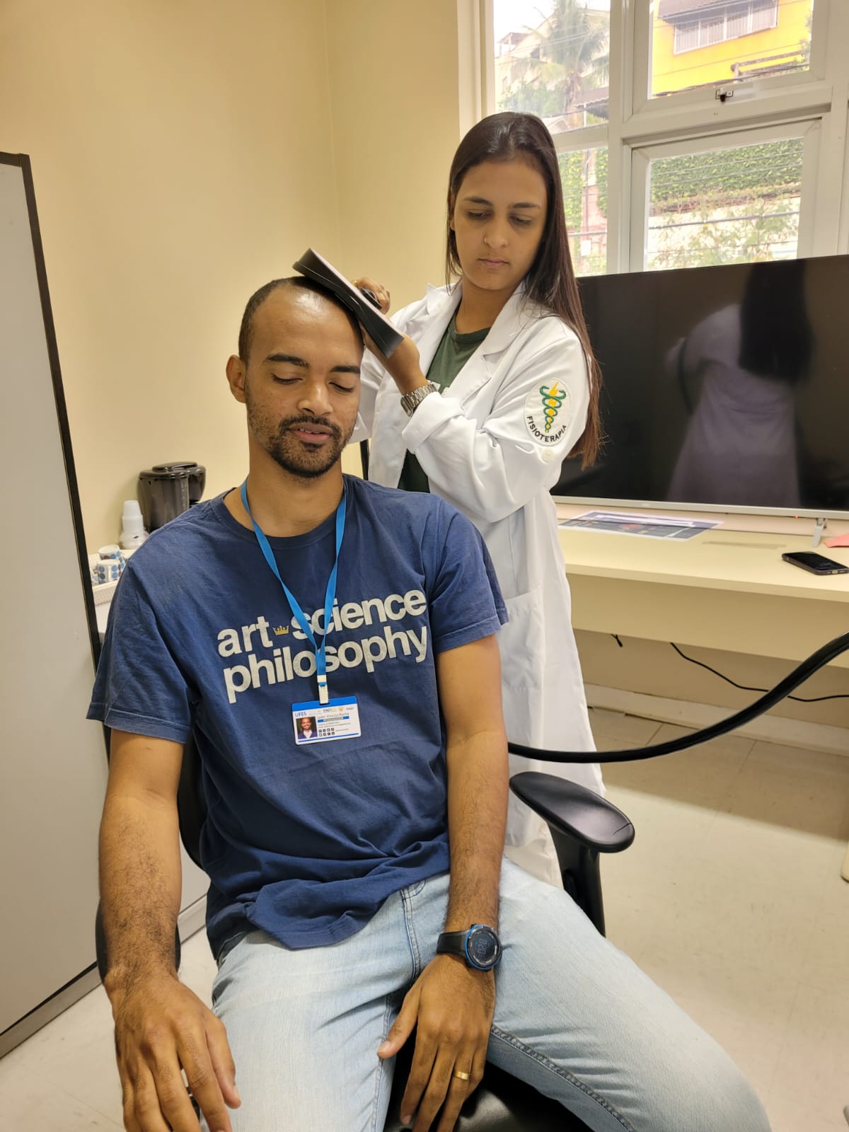 Foto de um homem sentado, sendo atendido por uma profissional vestida com um jaleco branco que passa um sensor sobre sua cabeça