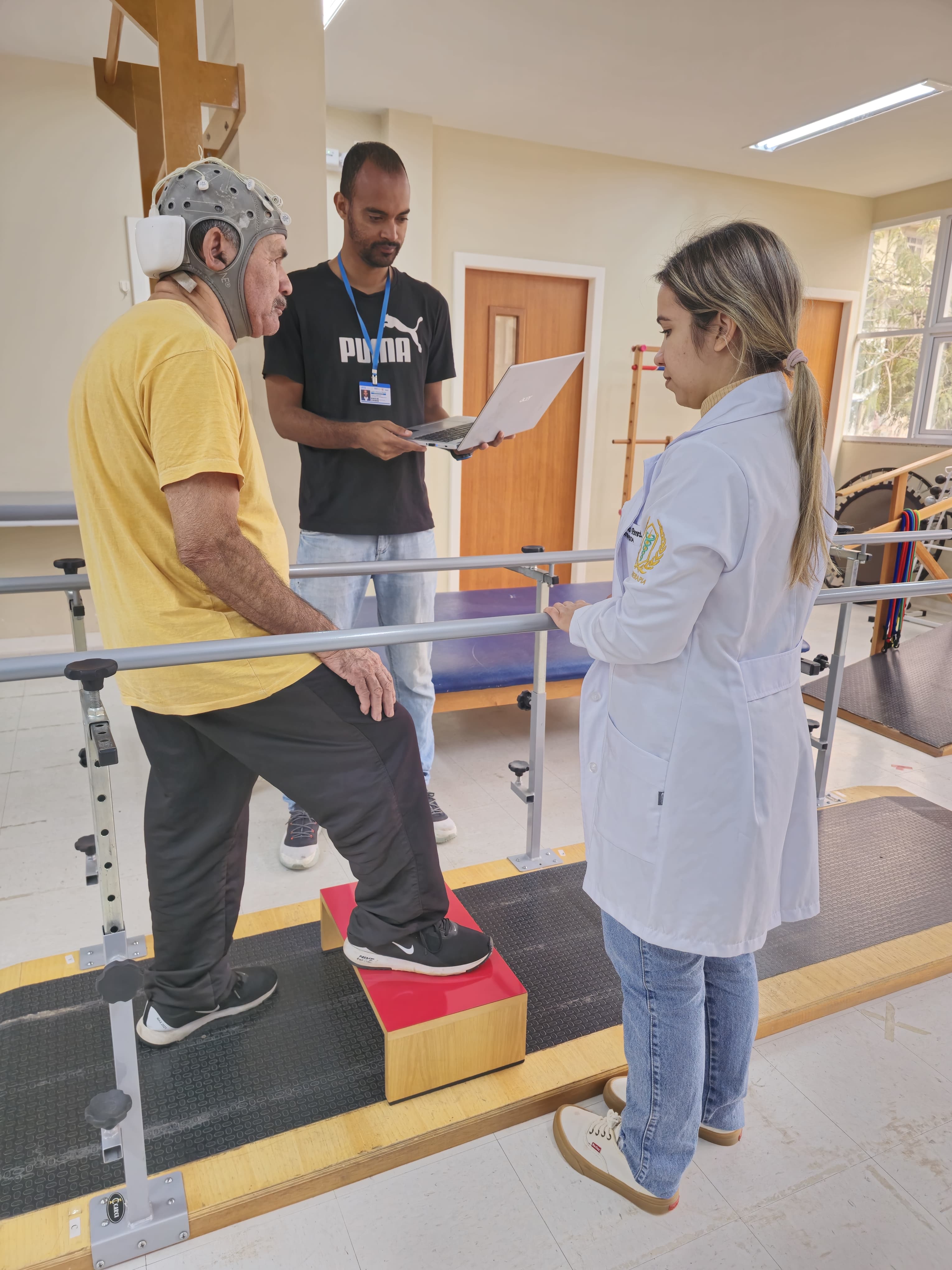 Foto de paciente em pé, com um pé sobre um caixote de madeira, sendo observado por dois profissionais