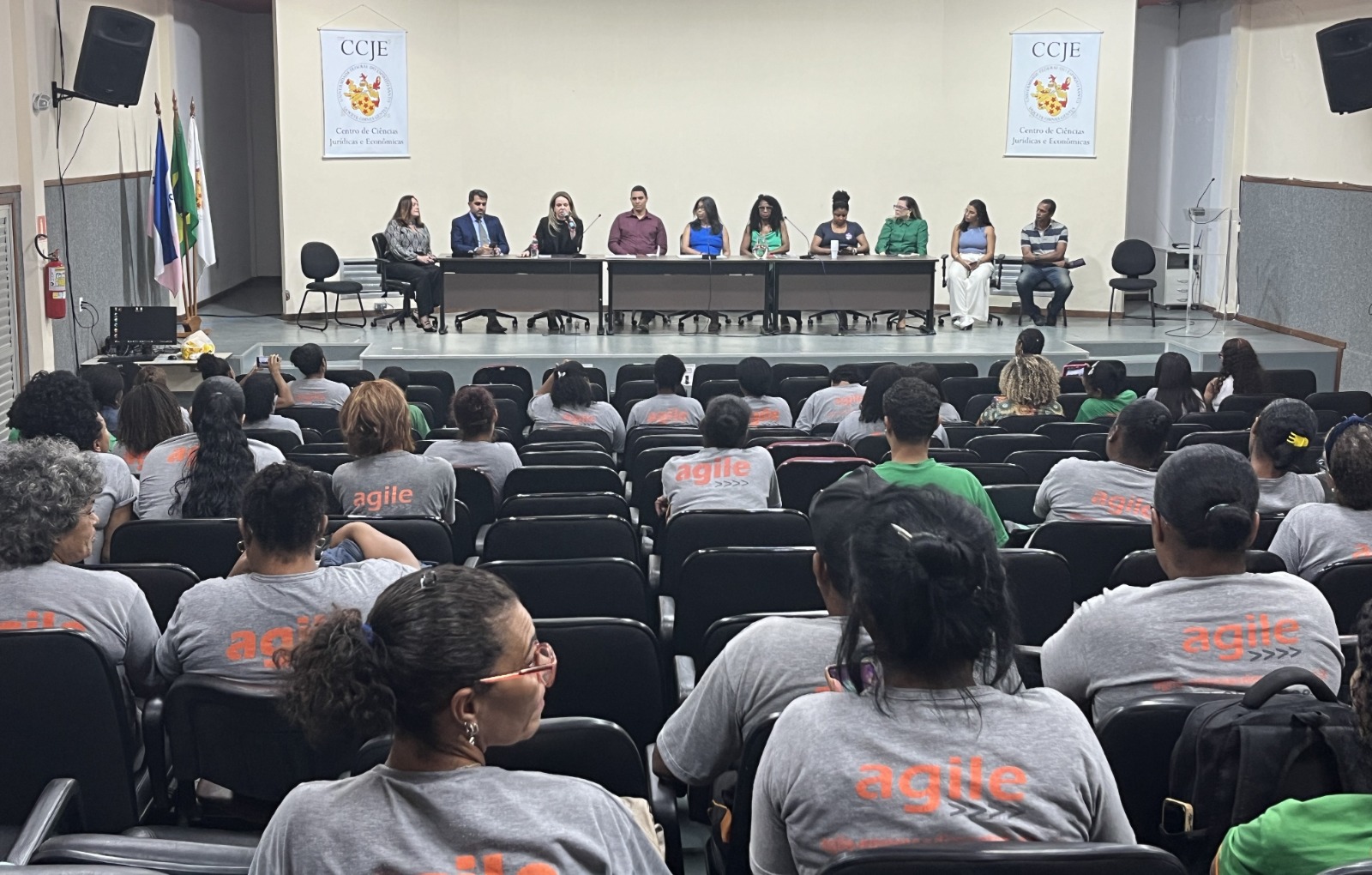 Foto do auditório com as trabalhadoras em primeiro plano (de costas) e a mesa coordenadora do evento ao fundo