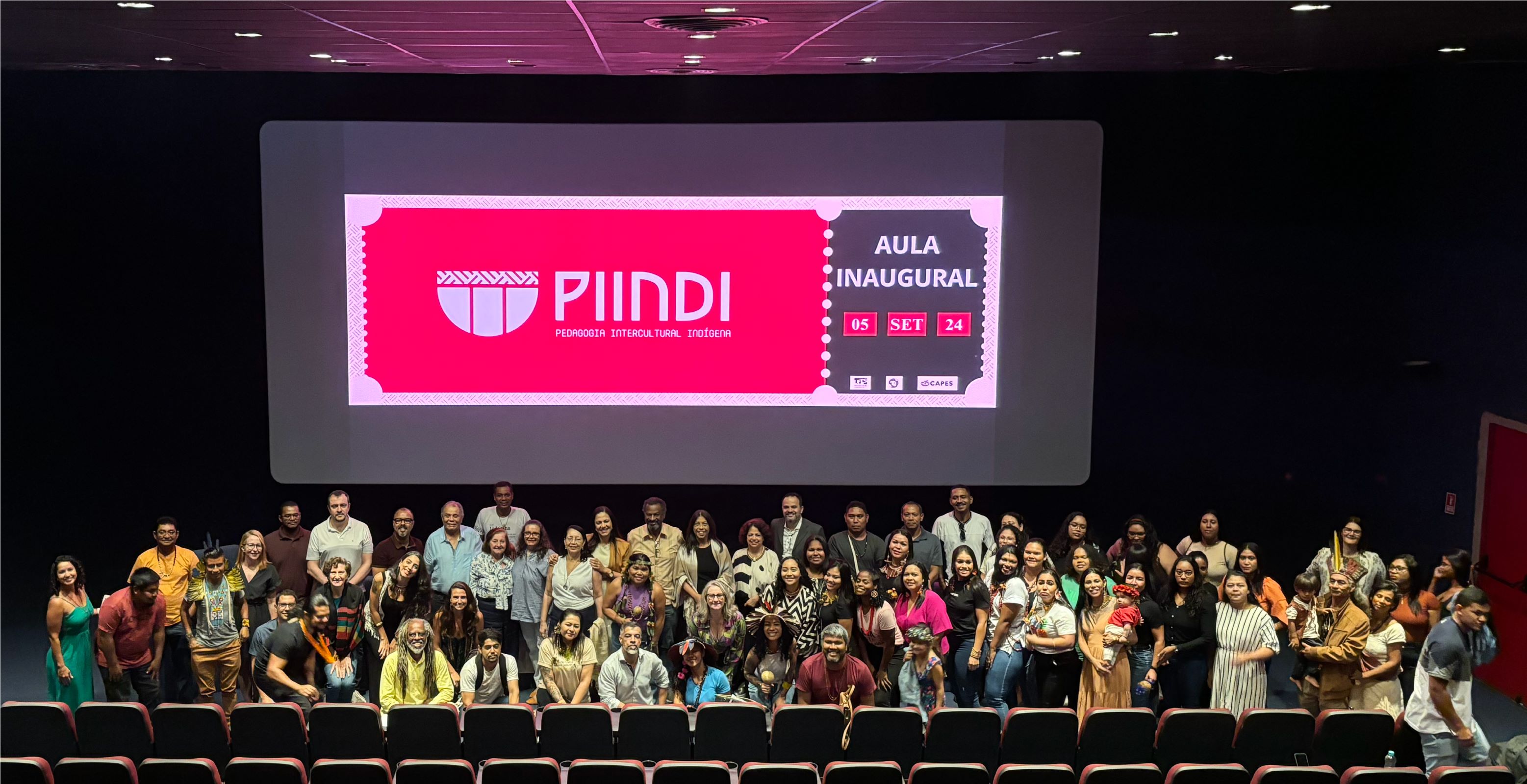 Foto da abertura da aula inaugural, com todos os presentes posando para a foto. Atrás, projetada na tela do cinema, a logomarca do curso e a data da aula.