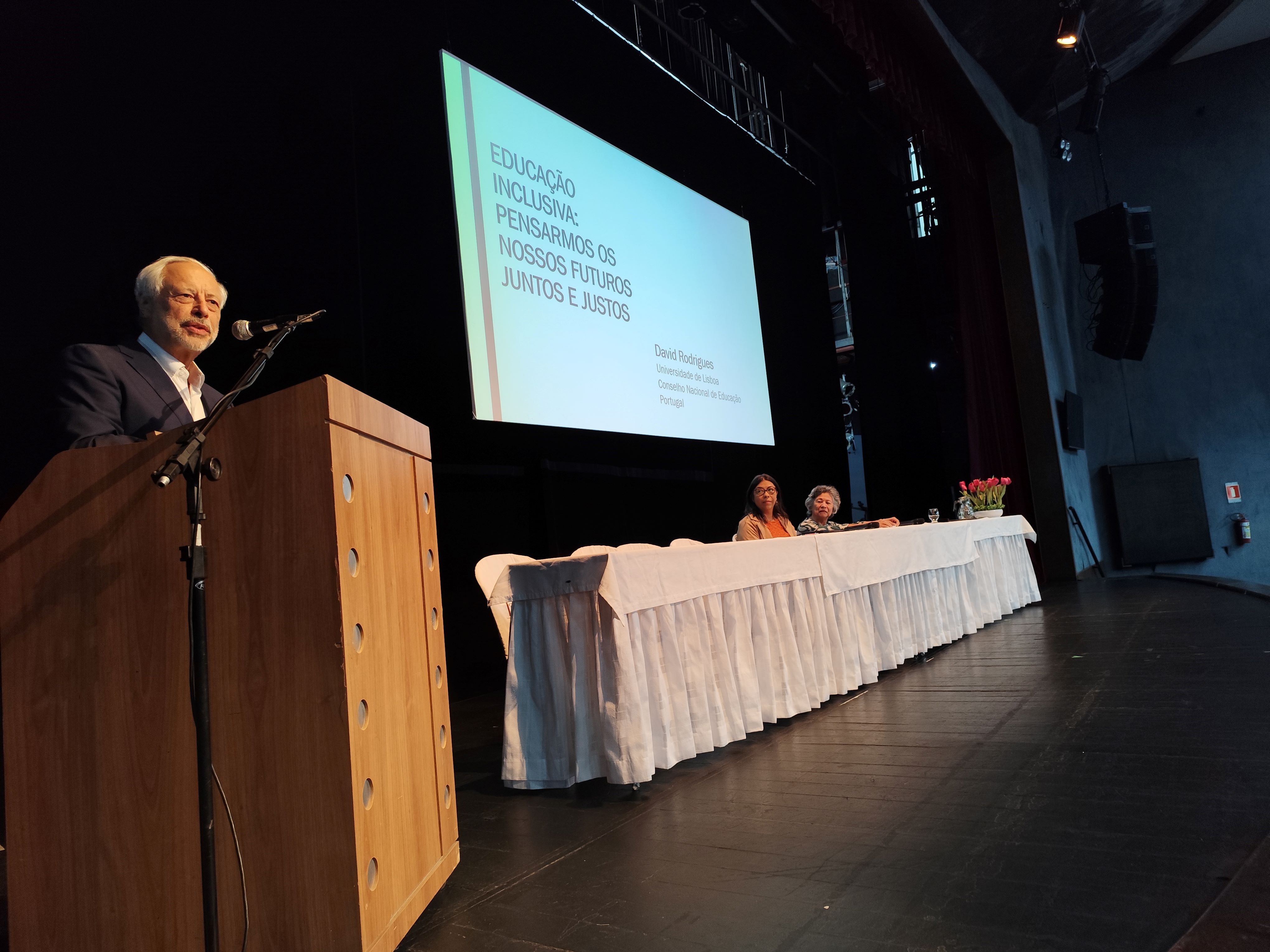 Foto do professor David Rodrigues, em primeiro plano, no púlpito do teatro. Ao fundo, a vice-reitora e a professora Denise Meyrelles sentadas à mesa principal