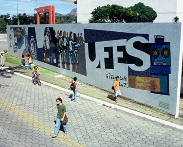 Foto onde mostra, do alto, o mural da Ufes e pessoas andando na calçada e atravessando a rua em frente ao mural