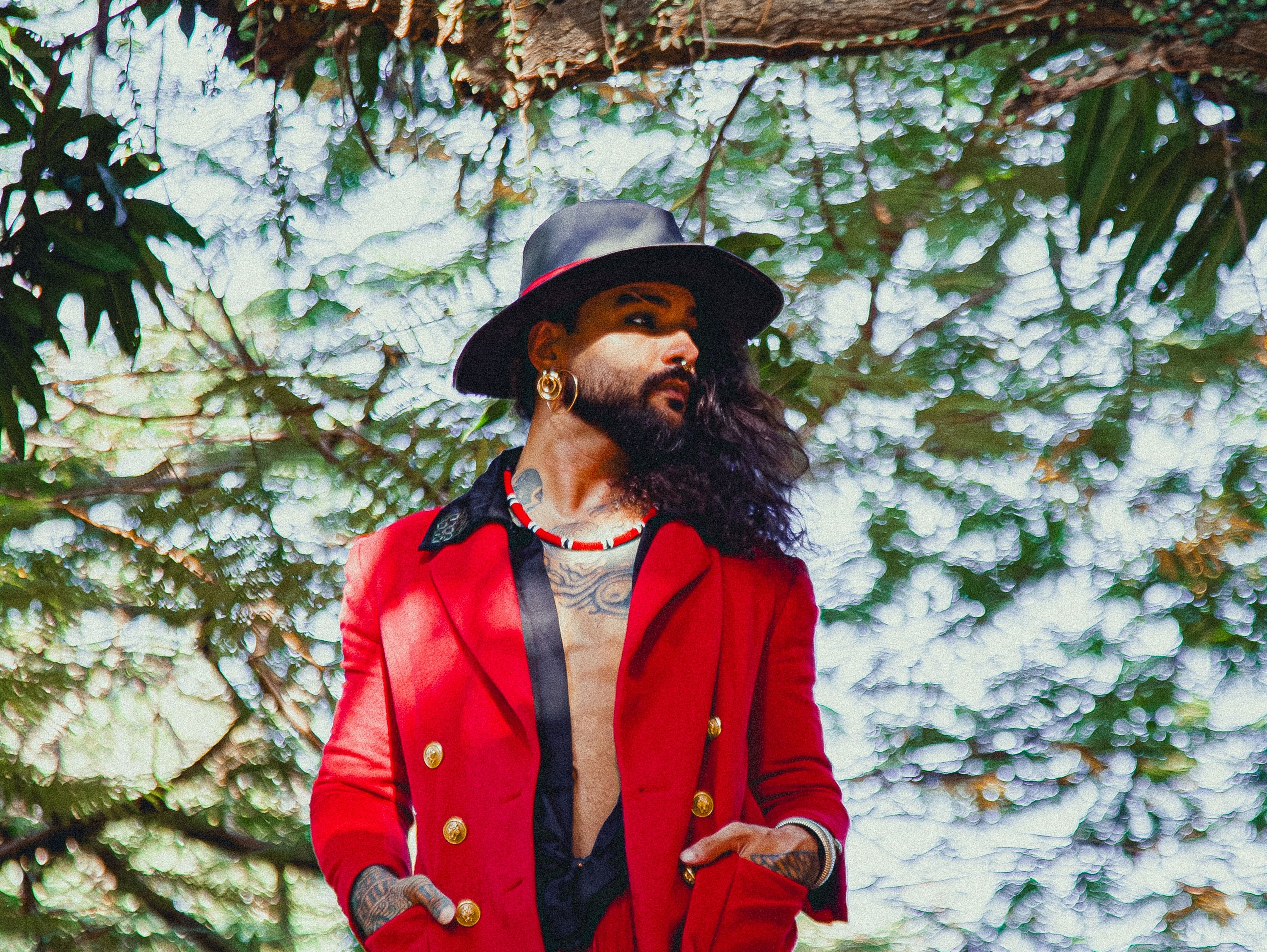 Foto do cantor e compositor André Prando. Ele está em pé, em um lugar com folhagens, veste um blazer vermelho sobre uma camisa preta e usa um chapéu preto.