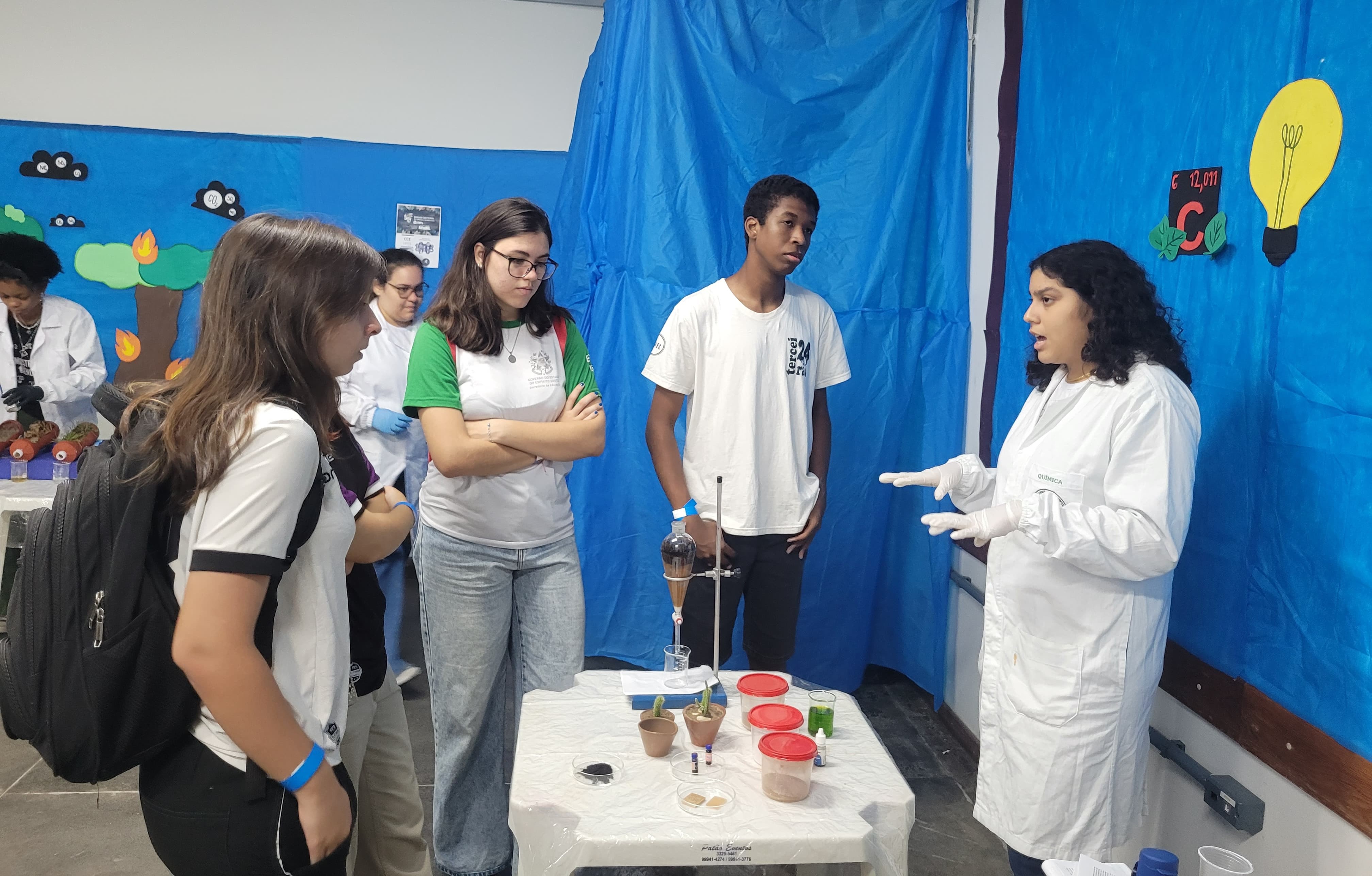 Foto de uma aluna da Ufes, vestindo jaleco branco, apresentando uma experiência para três estudantes visitantes