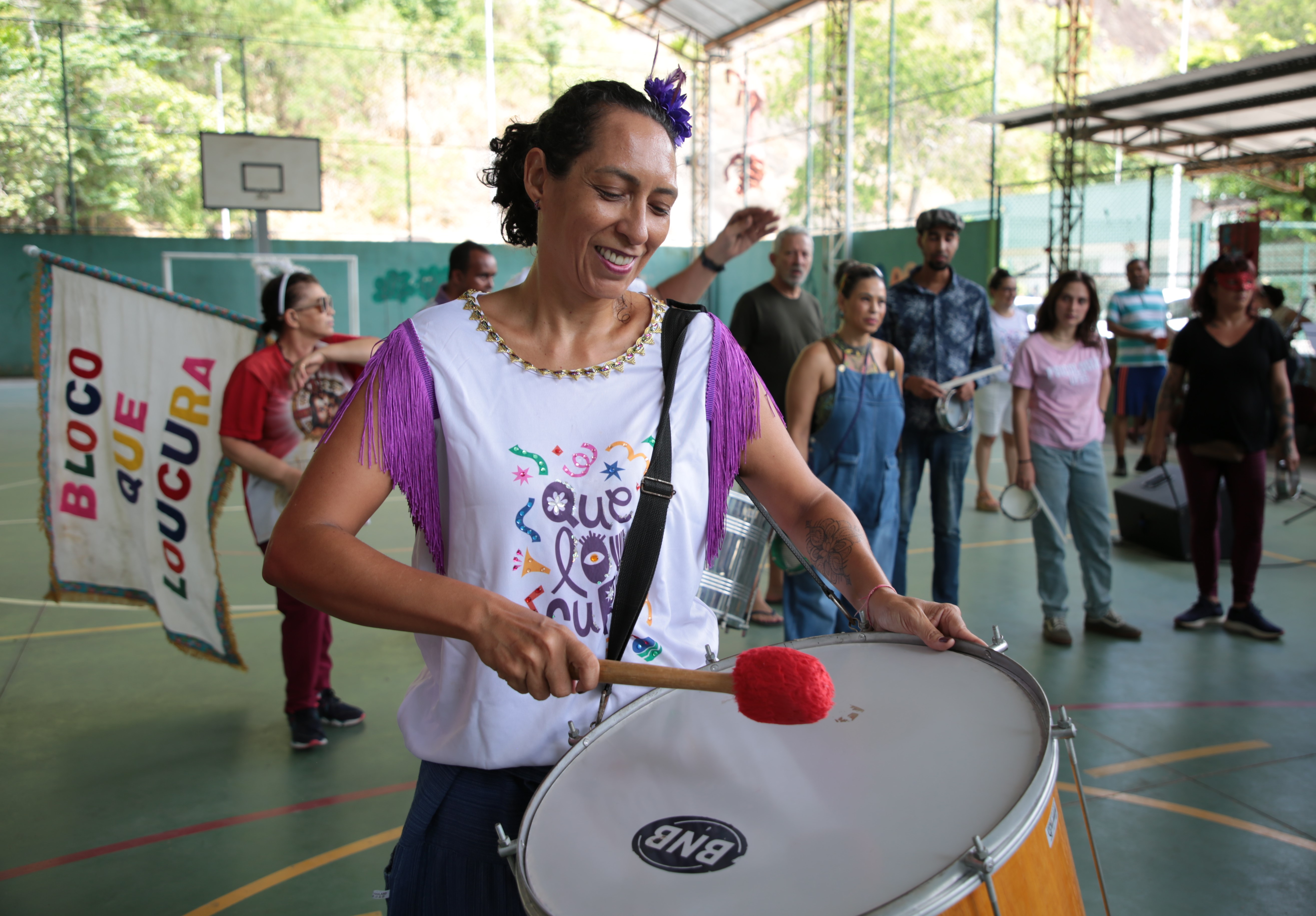 Foto de uma integrante do bloco tocando bumbo e outros participantes atrás