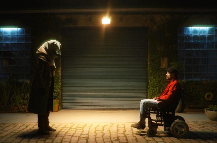 Cena de Memórias de um Esclerosado, na qual um homem em cadeira de rodas está de frente para uma pessoa em pé com uma cabeça de sapo