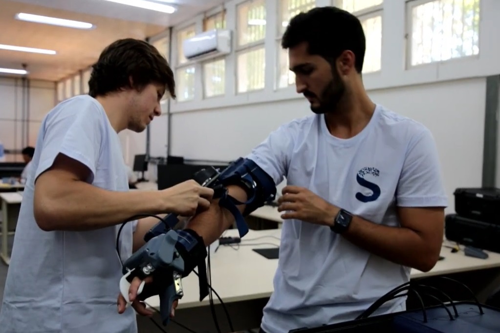 Foto de dois rapazes testando um braço mecânico desenvolvido dentro do Espaço