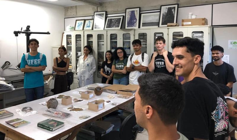 Foto de estudantes em uma sala interior do herbário onde há plantas expostas sobre uma mesa