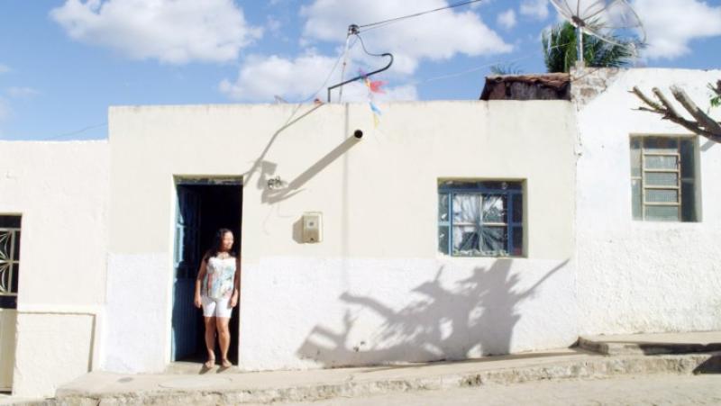 Cena de O Bem Virá, na qual aparece uma mulher encostada na entrada de uma casa branca