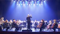 Foto da orquestra sinfônica com o maestro de costas regendo os músicos a sua frente