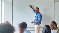 Imagem de um professor negro, de frente para um quadro branco, em uma sala de aula de jovens