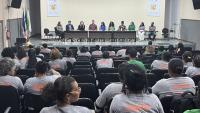 Foto do auditório com as trabalhadoras em primeiro plano (de costas) e a mesa coordenadora do evento ao fundo