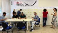 Grupo de participantes em uma mesa elaborando o planejamento enquanto outras pessoas fixam papeis na parede como parte da atividade