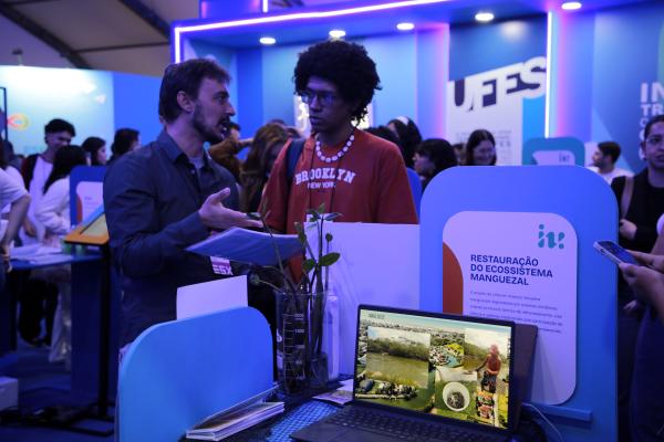 Foto de um professor apresentando um projeto a um visitante, na área interna do estande