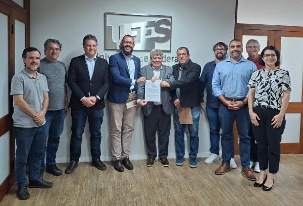 Foto dos participantes da reunião em frente à logomarca da Ufes, mostrando o acordo assinado