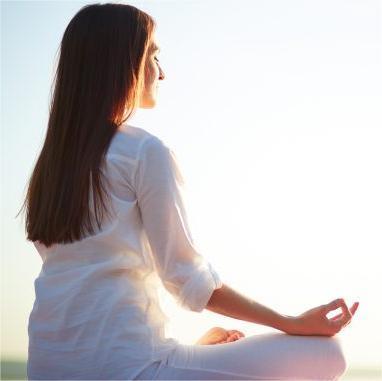 Foto de mulher branca, com cabelo longos, lisos e castanhos, vestindo roupas brancas e sentada em postura de meditação
