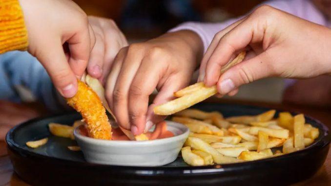 Foto de quatro mãos de pessoas diferentes passando batatas fritas num pote de molho.