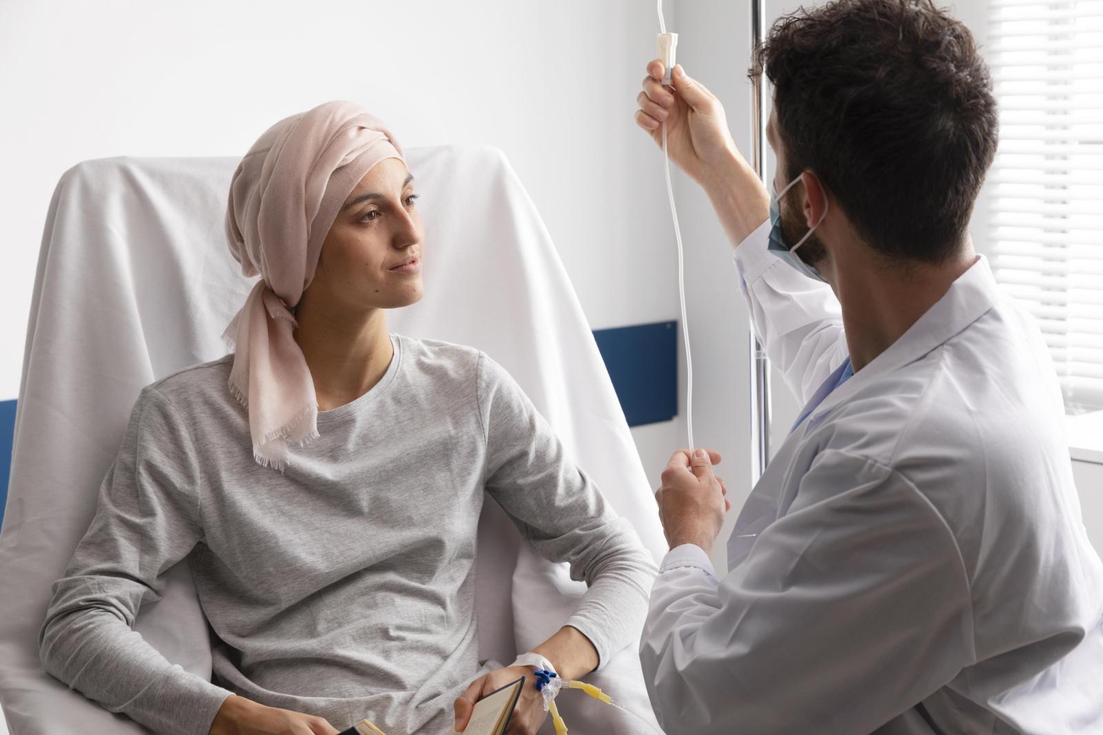 Imagem de uma mulher sentada em uma cama hospitalar com um lenço na cabeça e um enfermeiro ajustando a medicação no soro