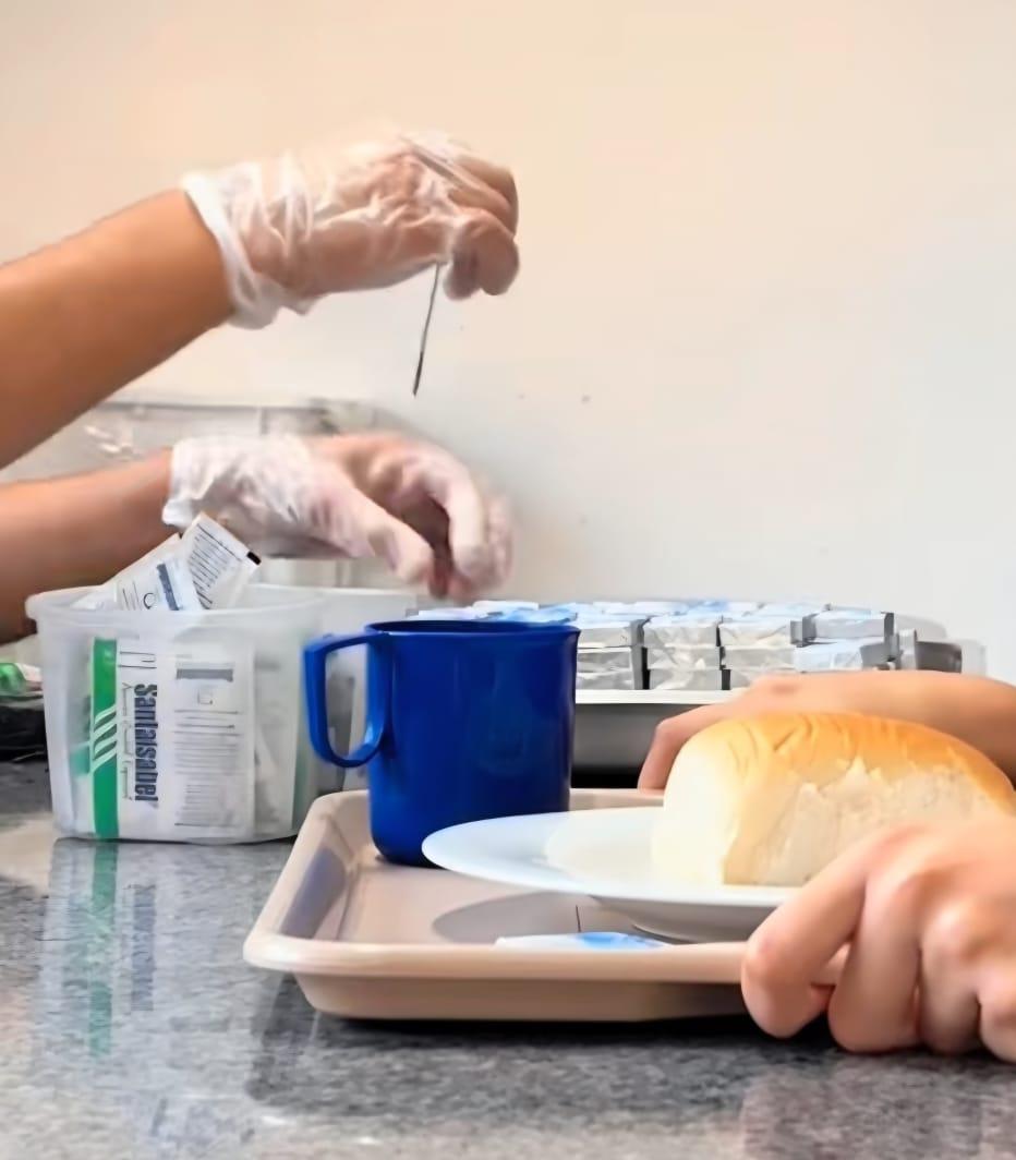 Foto de mãos segurando uma bandeja onde há um prato com um pão e uma xícara. Na imagem aparecem ainda duas mãos colocando um sachê de açúcar sobre a bandeja