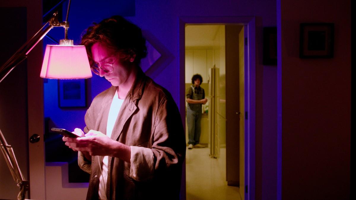 Cena de Labirinto dos Garotos Perdidos em que aparece um homem digitando no celular em uma sala com baixa iluminação enquanto um jovem observa de longe
