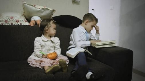 Cena de A Voz de Deus, na qual aparecem um menino sentado no sofá, lendo um livro. Ao seu lado, sua irmã o observa